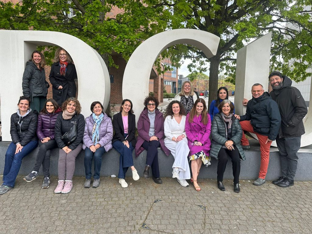 13 dones i 2 homes davant les lletres de formigó verticals DCU. La majoria estan assegudes a davant a sobre del socle de formigó; dues estan dretes a l'esquerra dins la D i dues assegudes al mig a segona fila dins la C. A la dreta, els dos homes estan drets davant de la U.